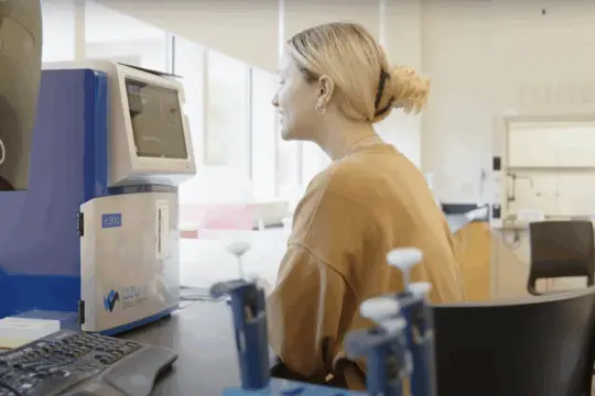 A person with blonde hair is sitting and smiling at a laboratory desk, looking at a device labeled "Azure Biosystems." The room is well-lit with windows in the background, and various lab equipment is on the table.